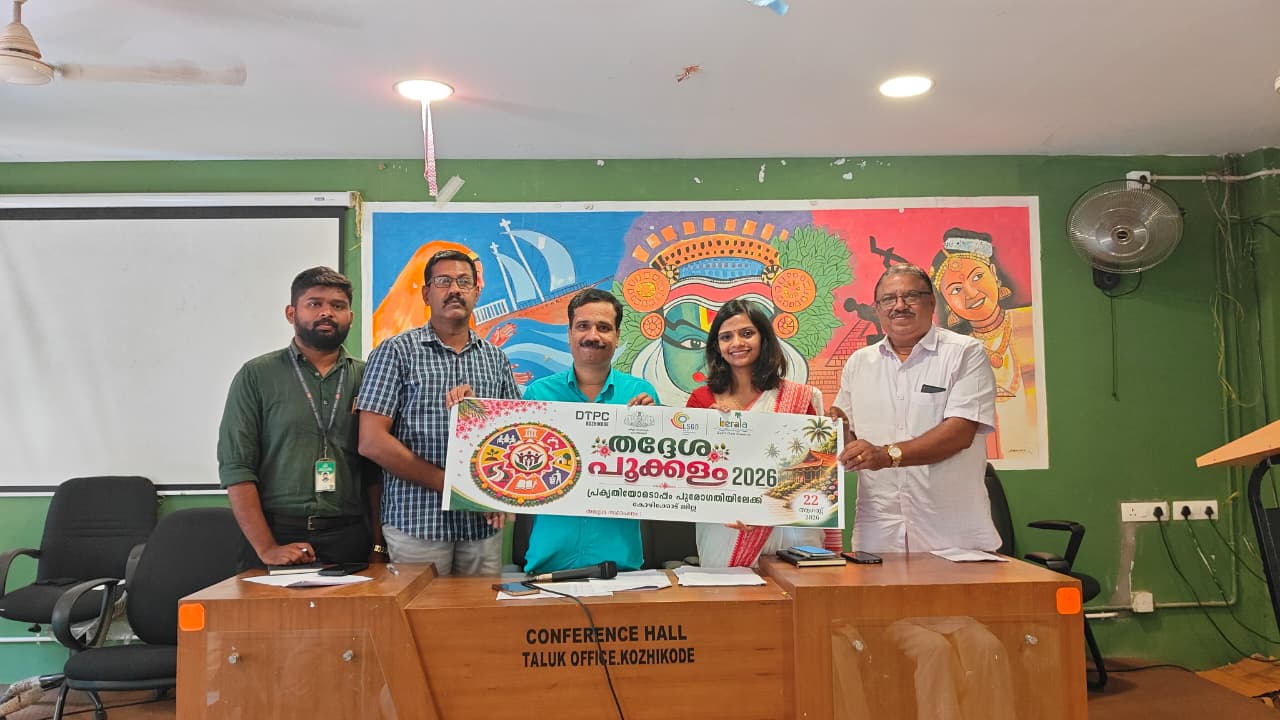 Five organisers standing behind a table in the Conference Hall of the Taluk Office, Kozhikode, holding up a wide Thadhesha Pookalam 2026 poster between them, in front of a painted mural of the city's landmarks and a Kathakali face